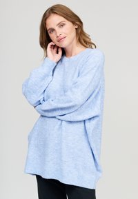 Hellblaues, übergroßes Sweatshirt, gestrickte Textur, Bootsausschnitt, gerippte Bündchen und Saum, weicher Abschluss. Kombiniert mit dunklen Hosen.