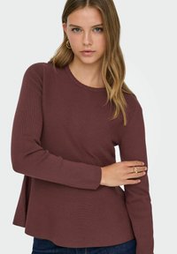 Bordeauxrotes Langarmoberteil mit gerippter Textur und Rundhalsausschnitt. Es verfügt über einen ausgestellten Saum und eng anliegende Ärmel. Kombiniert mit blauen Jeans.