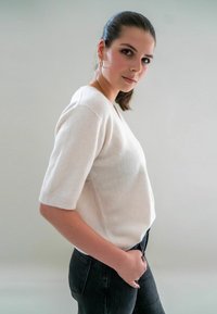 Pull blanc à manches courtes avec une coupe décontractée, fabriqué en tissu doux. Associé à un jean noir ajusté. Modèle se tenant de profil devant un arrière-plan neutre.