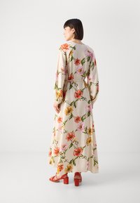 Maxi abito color crema con un vivace motivo floreale in rosso, rosa e giallo. Maniche lunghe e vita cintrata. Indossato con sandali con tacco a blocco rossi.