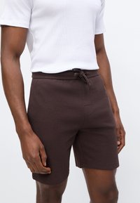 Άνδρας που φοράει σκούρα καφέ υφασμάτινη βερμούδα με κορδόνι στη μέση και λευκό κοντομάνικο πουκάμισο σε απλό φόντο.