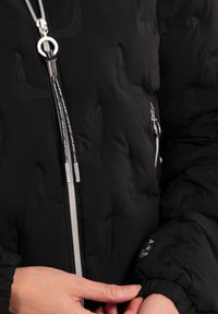 Schwarze Pufferjacke mit texturiertem Finish, ausgestattet mit einem zentralen Reißverschluss und elastischen Bündchen. Silberne Hardware-Akzente und Branding-Details sind sichtbar.