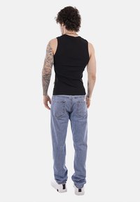 Uomo con braccio tatuato che indossa una maglietta nera senza maniche, jeans blu e sneakers bianche, in piedi di schiena su uno sfondo bianco.