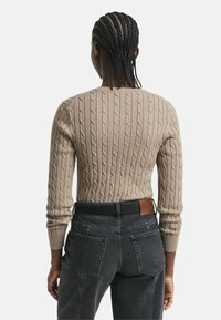Beige Kabelstrickpullover mit rundem Ausschnitt und langen Ärmeln, kombiniert mit dunkelgrauen hochgeschnittenen Jeans. Texturiertes Muster auf dem Pullover sichtbar.