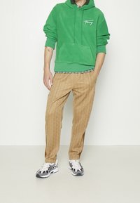 Felpa verde in poliestere con tasca a marsupio e logo; pantaloni marroni con motivo e spacchi laterali; scarpe sportive black and white con dettagli.