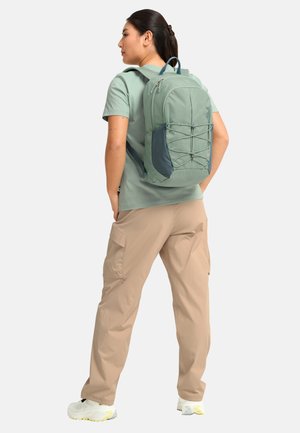 Frau trägt ein hellgrünes T-Shirt und beige Cargohosen, steht mit grünem und grauem Rucksack nach links gewandt, Hände in den Taschen.