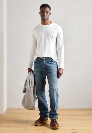 White long-sleeve shirt, blue jeans with faded pattern, brown shoes with thick soles. Gray hoodie draped over arm. Wooden floor backdrop.