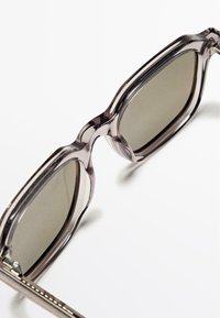 Okulary przeciwsłoneczne z szarymi szkłami i przezroczystą, grubą plastikową oprawą. Oprawa ma zaokrąglony kształt i subtelne akcenty stylistyczne na zausznikach.