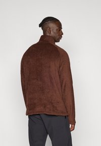 Brauner, strukturierten Fleece-Pullover mit hohem Kragen, der einen entspannten Schnitt und ein geripptes Muster aufweist, von hinten mit dunklen Hosen gezeigt.