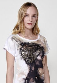 T-shirt blanc à manches courtes avec un imprimé artistique noir et doré représentant le profil d'une femme, avec un motif stylisé sur sa tête.