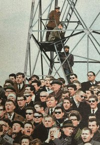Menigte van mannen in verschillende outfits, waaronder pakken en hoeden, sommigen dragen zonnebrillen. Drie individuen op een metalen structuur op de achtergrond.
