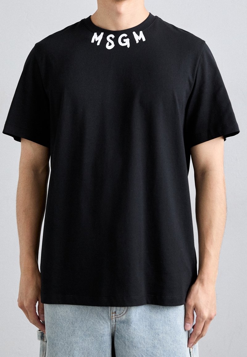 Homme portant un T-shirt noir à manches courtes avec les lettres blanches "MSGM" près du col, assorti à un short en denim bleu clair.