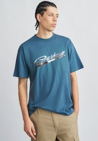 Camiseta de algodón azul con un logo metálico plateado de "Carhartt". Mangas cortas, cuello redondo, ajuste relajado, textura suave.