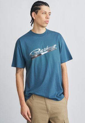 Camiseta de algodón azul con un logo metálico plateado de "Carhartt". Mangas cortas, cuello redondo, ajuste relajado, textura suave.