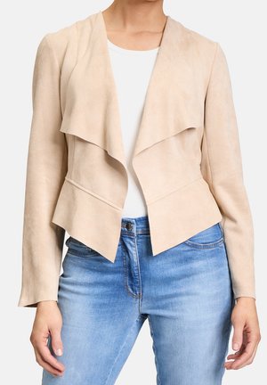 Femme portant une veste beige en cuir suédé ouverte sur un haut blanc et un jean bleu clair, debout avec les mains près des hanches.