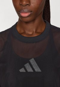 Zwarte sporttop met een transparant ontwerp, geribbelde halslijn en een prominent grijs-wit adidas-logo op de voorkant.