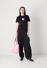 Black short-sleeve t-shirt with a colored logo, paired with loose black cargo pants. Carrying a pink tote bag with short handles and pointed heels.