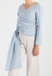Blouse cache-cœur rayée bleu clair et blanc, avec un décolleté en V profond, des manches longues et un détail noué à la taille. Portée avec un pantalon beige à jambes larges.