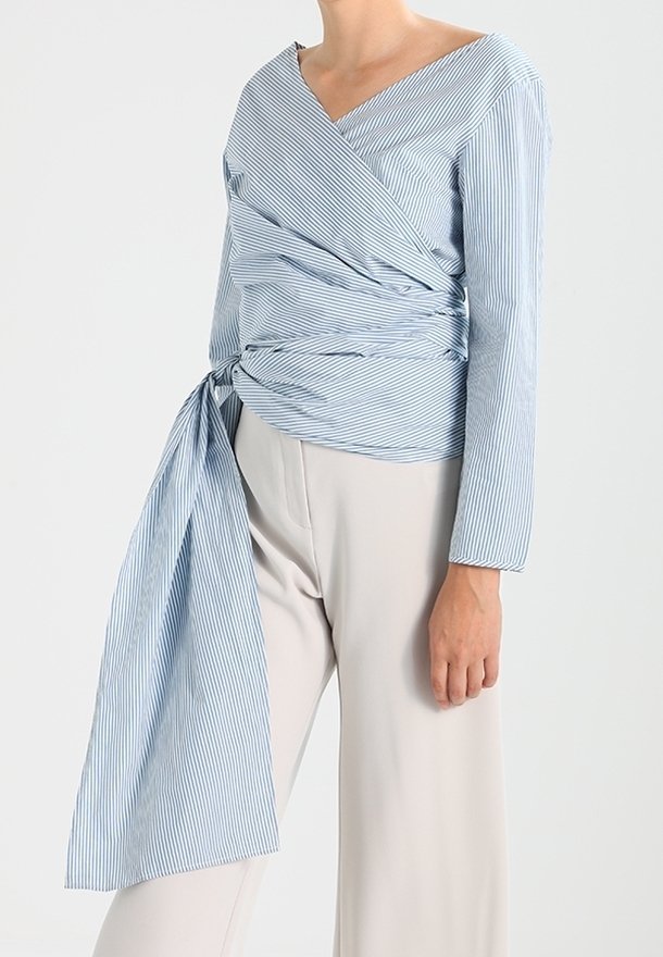 Blouse cache-cœur rayée bleu clair et blanc, avec un décolleté en V profond, des manches longues et un détail noué à la taille. Portée avec un pantalon beige à jambes larges.
