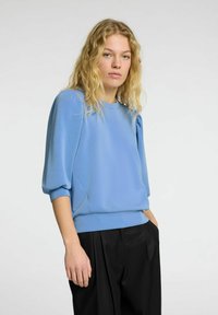 Gai zils džemperis ar trīs ceturtdaļu puff piedurknēm un apaļu kakla izgriezumu. Mīksta, gluda tekstūra. Saskaņots ar melniem, brīviem biksēm.