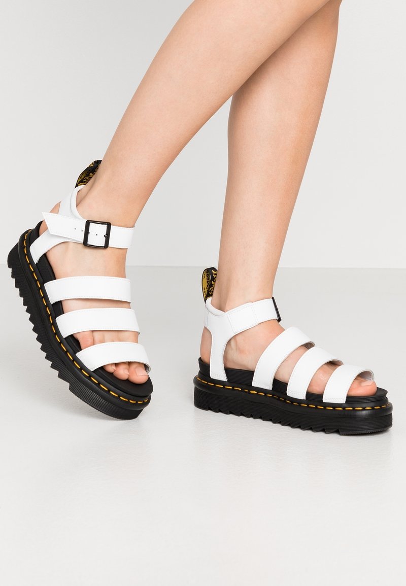 Jambes portant des sandales à plateforme en cuir blanc avec semelles noires et surpiqûres jaunes, comportant trois brides et des boucles à la cheville.