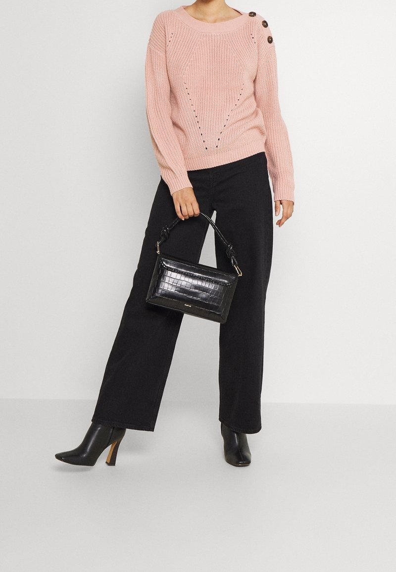 Pull rose clair en maille avec des trous décoratifs et des boutons sur le côté, associé à un pantalon large noir et un sac à main noir embossé effet crocodile.
