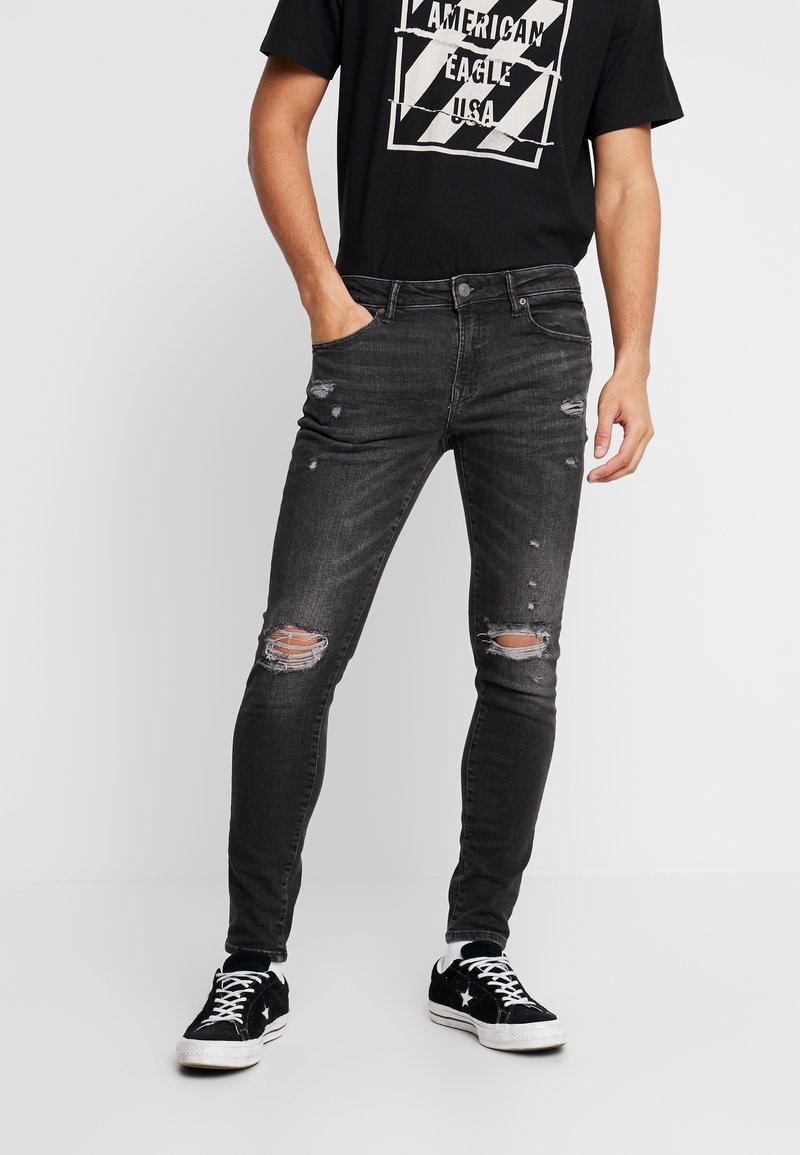 Jean skinny noirs avec des déchirures aux genoux, fabriqués en tissu denim. Stylisés avec un t-shirt graphique noir et des baskets noires.