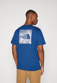 Zilā kokvilnas T-krekls ar īsām piedurknēm, ar baltu grafiku aizmugurē, kurā attēlotas viļņi un teksts "THE NORTH FACE".