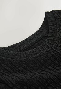 Mustast tekstureeritud kangast, millel on kortsus välimus, ümar kaelus ja soonikserv. Lähedalt vaadatuna rõhutab materjali mustrit.