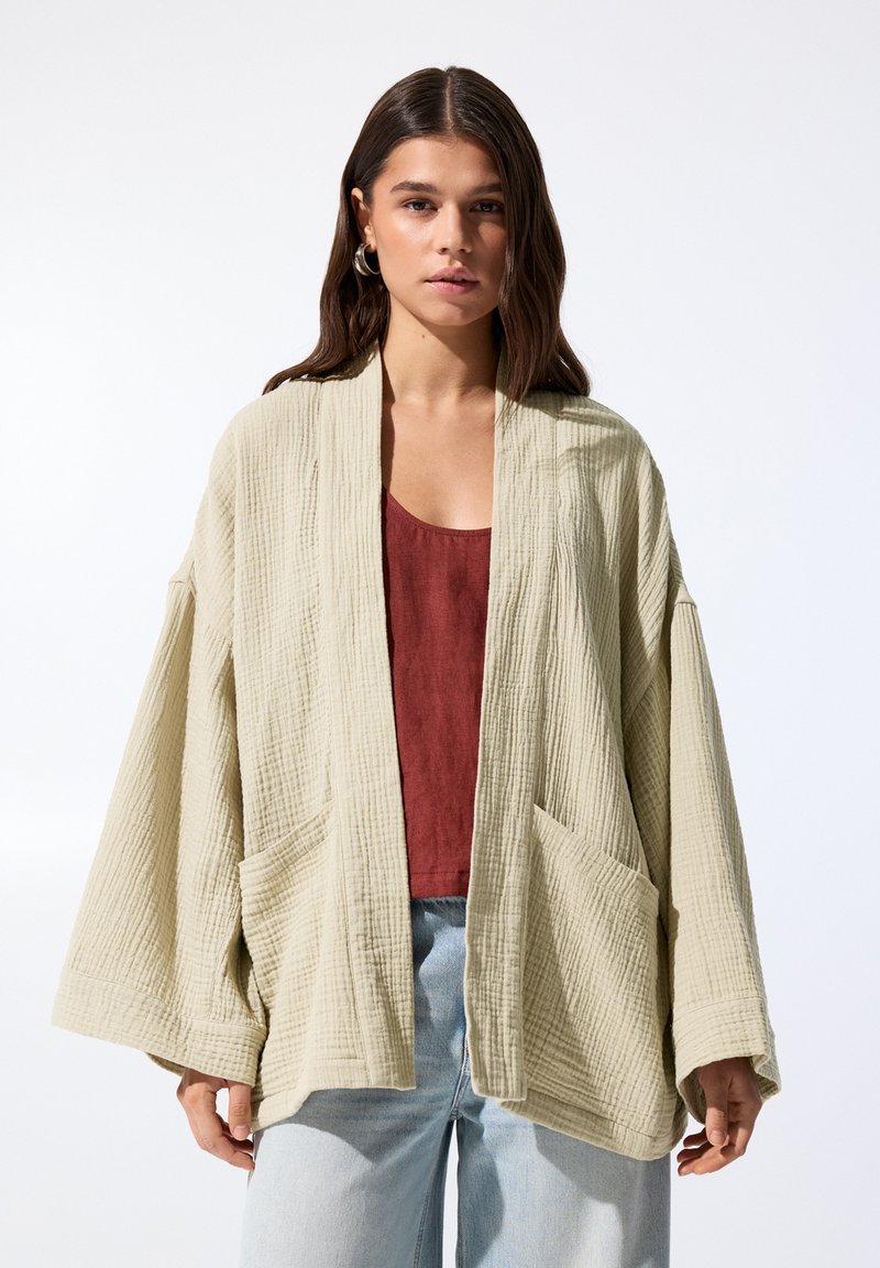Bežna teksturirana oversized jakna s sproščenim krojem, ki vključuje dva sprednja žepa in tričetrtinske rokave, oblečena čez maroon top.