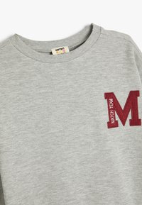 Grijze sweatshirt met een geribbelde ronde hals en een bordeauxrode "M" geborduurd op de linkerborst. Zachte, gestructureerde stof.