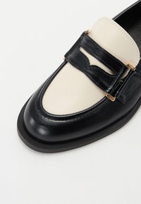 Svart og kremfarget lærloafers med rund tå, gulldetaljer og en teksturert rem over vrista. Flat såle.