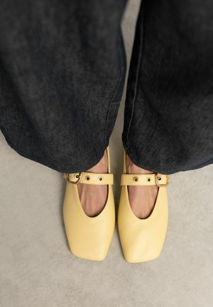 Voeten met gele Mary Jane-schoenen met een vierkante neus en gespsluitingen, gecombineerd met donkere wijde broekspijpen op een lichte vloer.