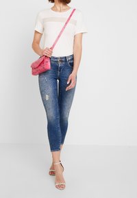 Top blanco de manga corta con panel transparente, jeans azules desgastados, bolso cruzado de patrón rosa y sandalias metálicas con tiras.