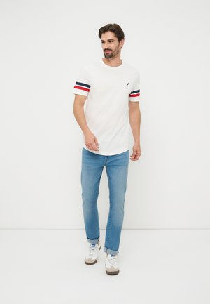 Man loopt, draagt wit T-shirt met rode en blauwe strepen op de mouwen, lichtblauwe spijkerbroek en witte sneakers tegen een effen witte achtergrond.