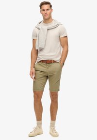 Beige t-shirt, olijfgroene shorts, lichtgrijze sweater over de schouders, bruine riem, crèmekleurige sneakers en lichtgrijze sokken.