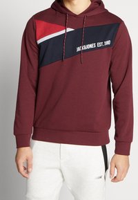 Sudadera de color burdeos con un diseño de bloques de color geométrico en rojo, marino y gris, capucha con cordones y lettering "JACK&JONES EST. 1990".