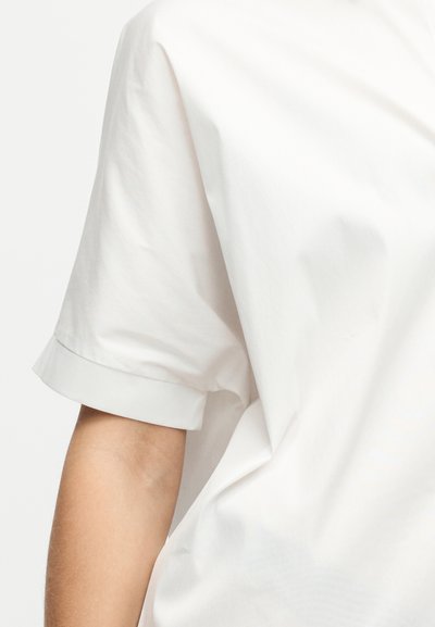 Chemise blanche à manches courtes avec un revers roulé sur le bras d'une personne, présentée sur un fond clair uni.