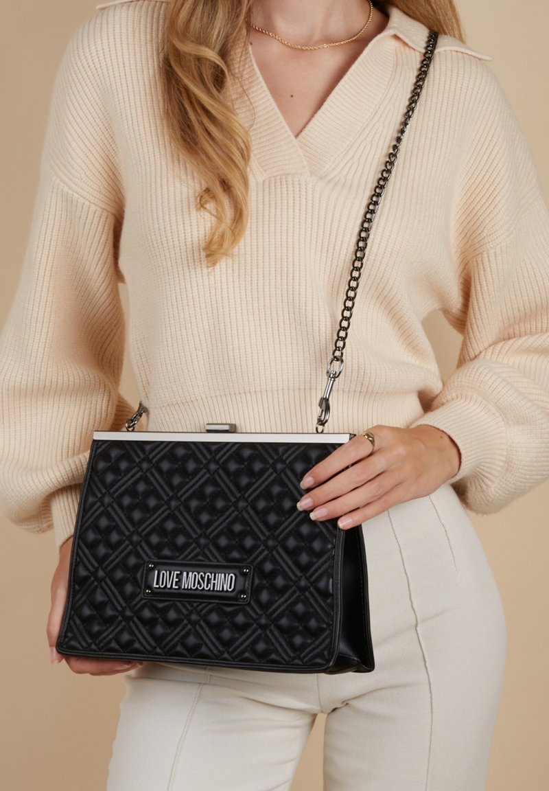 Schwarze gesteppte Handtasche mit einem silbernen Kettenriemen, rechteckiger Form, Metallverschluss und "LOVE MOSCHINO" Logo-Platte auf der Vorderseite.