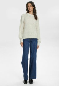 Cremfarbener, grob gestrickter Pullover mit gerippten Bündchen, kombiniert mit hochgeschnittenen blauen Jeans und schwarzen Stiefeletten. Einfaches, strukturiertes Design.