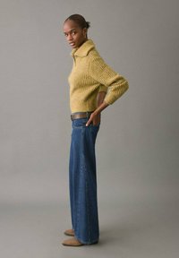 Jersey de punto trenzado mostaza con un cuello alto, combinado con unos vaqueros de tiro alto y acampanados azules. La modelo está de perfil con las manos en las caderas.