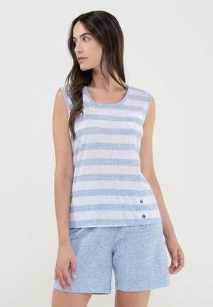 Mujer vestida con una blusa sin mangas blanca con rayas azules, combinada con pantalones cortos azul claro, de pie frente a un fondo liso y claro.