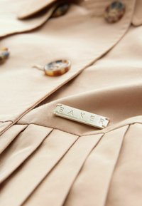 Beige stofjakke med dekorative knapper og et guldtonet metalmærke med indskriften "BAKER", med folder nær kanten.