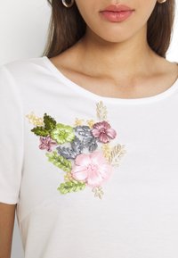 Biała koszulka z haftowanym wzorem kwiatowym w kolorach różowym, zielonym, fioletowym i szarym, z złotymi akcentami oraz delikatnymi wzorami liści.