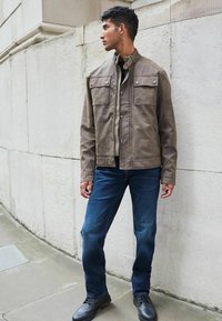 Chaqueta de cuero marrón con dos bolsillos en el pecho, botones plateados y un cuello; combinada con jeans azul oscuro y botas negras hasta el tobillo.