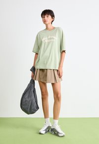 T-shirt verte oversize avec l'impression "New Balance Athletics", shorts plissés beiges, sac à bandoulière gris, baskets blanches et chaussettes blanches.
