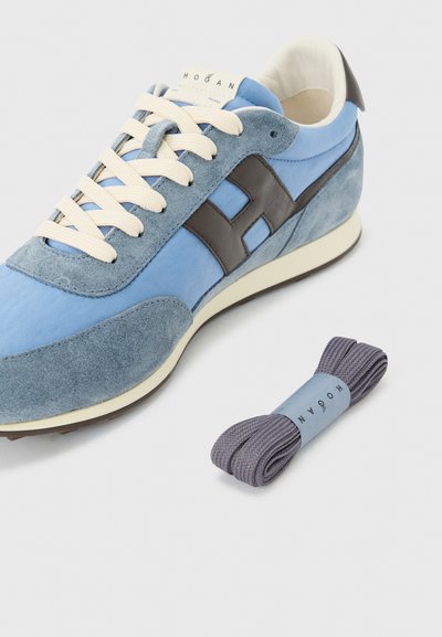 Baskets en daim bleu et gris avec des lacets crème, un logo latéral noir, et une paire supplémentaire de lacets gris à côté, sur un fond blanc.