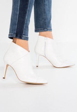 Bottes blanches en cuir à talons hauts et bouts pointus, avec fermetures éclair latérales, portées avec un jean court bleu foncé.