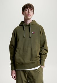 Olivenfarbener Kapuzenpullover aus Baumwollmischung mit einer frontalen Tasche und einem kleinen Logopatch. Rippbündchen und Saum.
