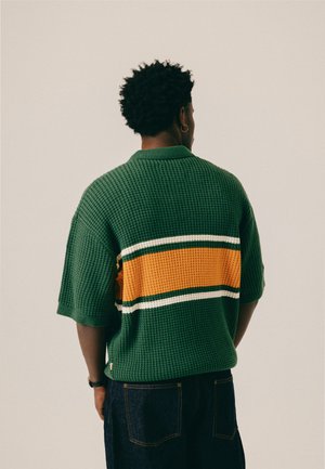 Mand med kort krøllet hår og guldring i øret iført grøn tekstureret kortærmet sweater med orange og hvid stribe på tværs af ryggen.